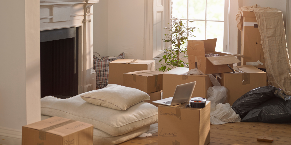 A cluttered home with moving boxes, coffee on the floor, and a laptop sitting on a box.