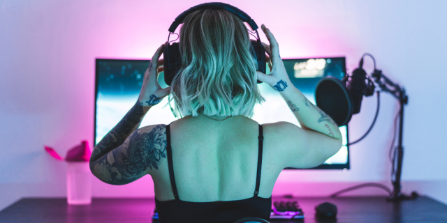 A woman with blonde shoulder-length hair puts on headphones, about to start a gaming livestream in front of her monitor which is flowing in bisexual lighting.