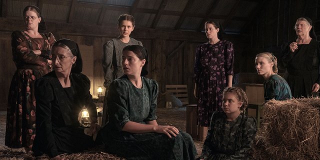 The primary cast of Women Talking in traditional Mennonite garb sit in a barn, all looking concerned in the same direction.
