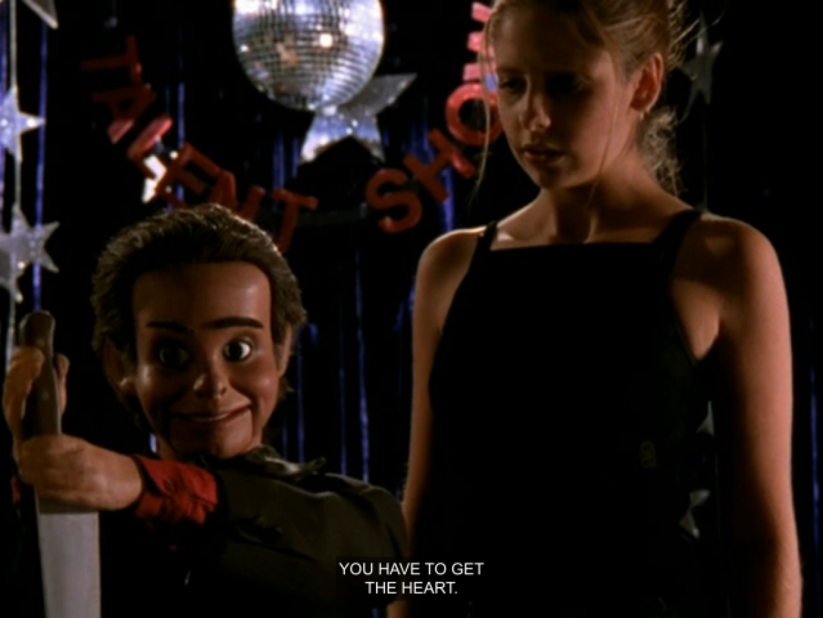 Buffy wearing a black spaghetti strap dress, standing on stage with a ventriloquist dummy who is about to plunge a knife into a demon. The dummy is saying “You have to get the heart.”