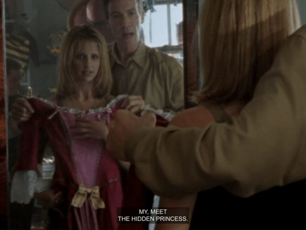 Ethan Rayne holding up a pink corseted dress for Buffy to see in the mirror. He’s saying “my, meet the hidden princess.