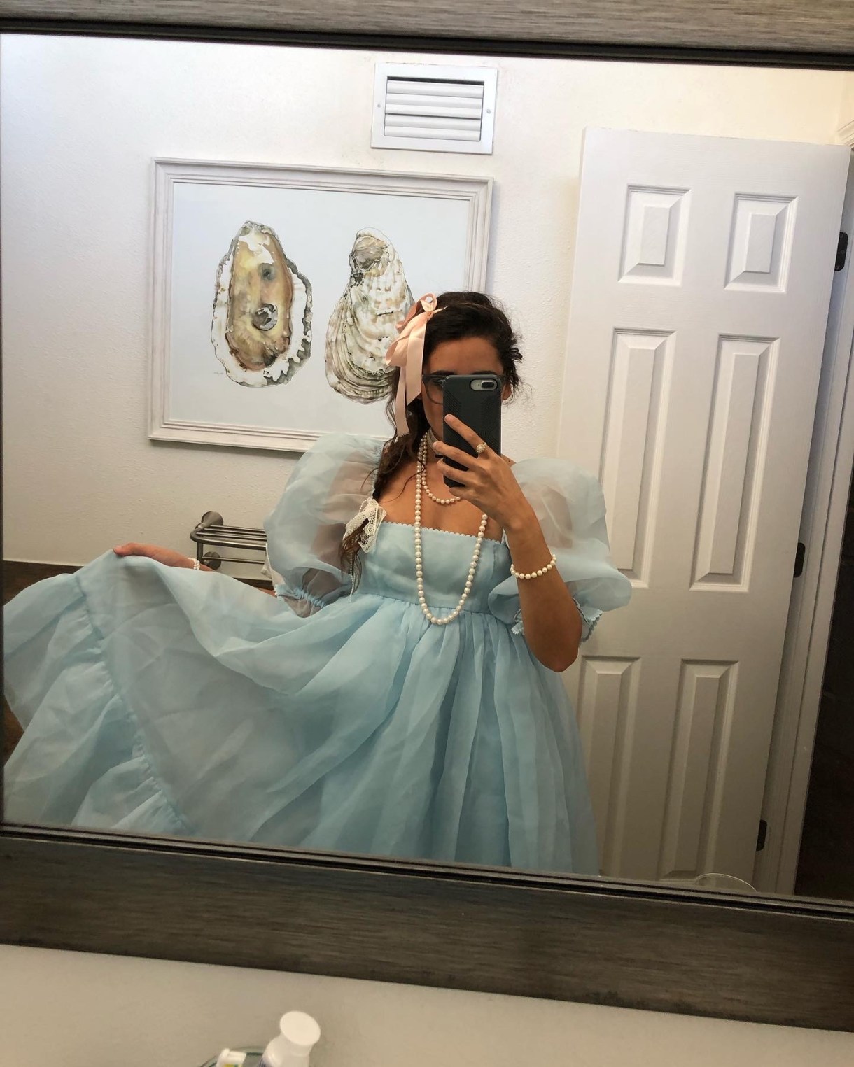 Kayla Kumari Upadhyaya wears a blue puffy dress, a pink ribbon in her braided hair, and several pearl necklaces. An oyster painting is behind her.