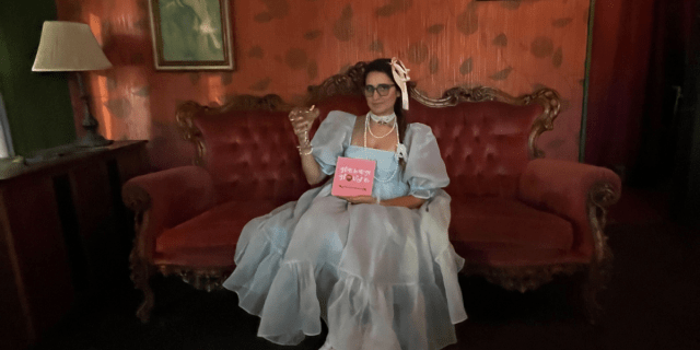 Kayla sits on an ornate Victorian sofa holding aloft a cocktail in a stemmed glass. She is wearing a doll-esque baby blue dress that is all gauzy with incredibly puffy sleeves, layers of pearls and a lace choker, ribbons in her hair which is braided, and glasses. She is holding her book, Helen House, which is pink with the title on it in white whimsical font and the "o" in house replaced with a smudgey lipstick kiss.