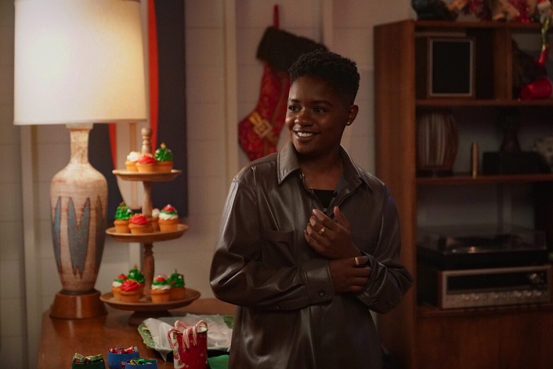 Coop, wearing a black shirt and brown leather jacket, stops by the boys' Christmas party. She is standing in front of a tower of Christmas cupcakes.