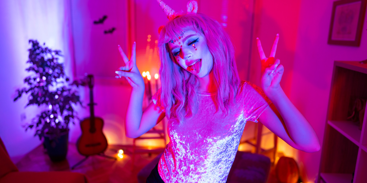 An Asian girl in white face pant and sparkles, dressed in pink velvet and a pink wig with unicorn ears gives peace signs and sticks her tongue out at the camera.