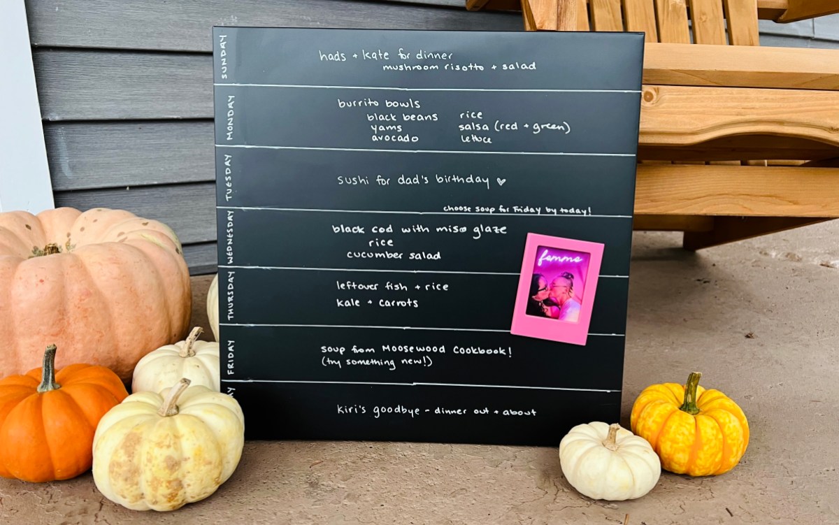 a chalkboard broken up into days of the week with a meal for each day sits on a porch, with small pumpkins on either side and an adirondack chair in the background. pinned to the chalkboard is a pink-tinted photo of two people kissing, with "femme" written across the top in neon letters.