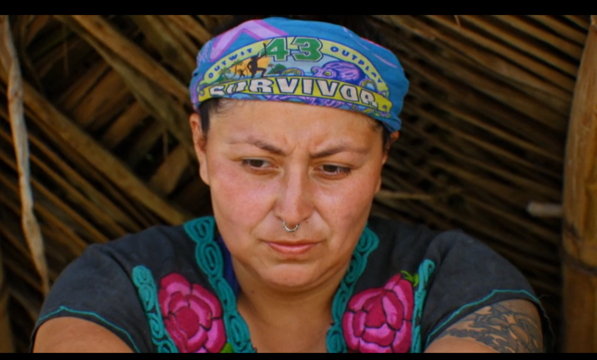 Karla Cruz Godoy in the reality TV show Survivor, wearing a blue buff and a colorful shirt, looks deep in thought