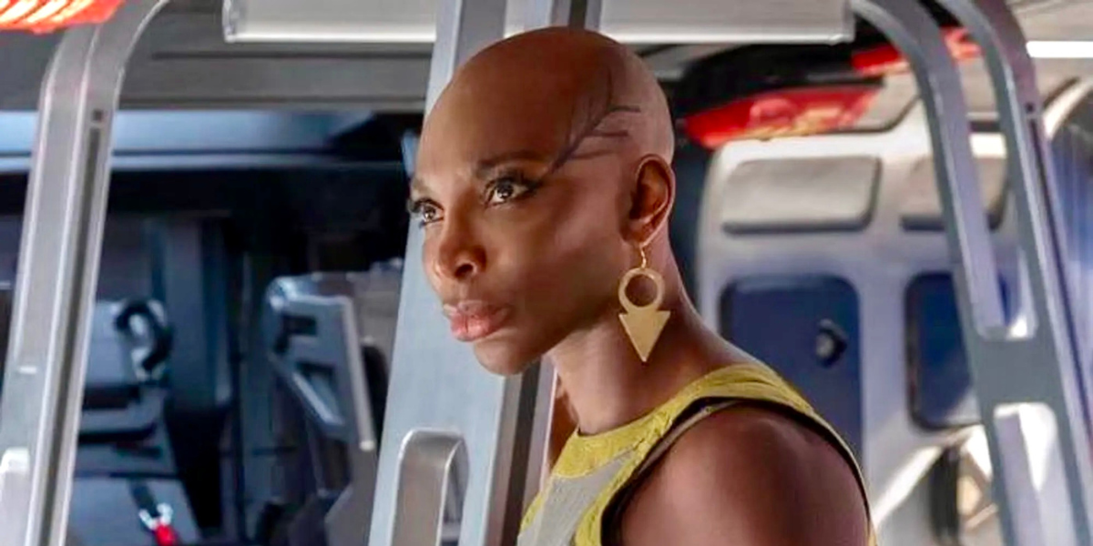 Michaela Cole as Aneka in Black Panther: Wakanda Forever, she looks to the right of camera and her hair is shaved bald. She is wearing big gold earrings and has detailed eye makeup.