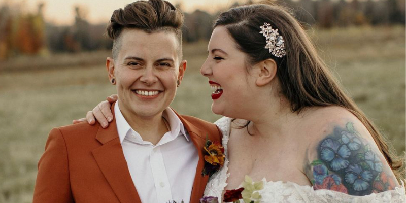 Mary Lambert is in a wedding dress that off the shoulder cuffs and her tattoo sleeve showing, she is facing her partner Wyatt, who has short cropped hair and is wearing an orange suit with a white dress shirt, and smiling. They are in front of a fall field.