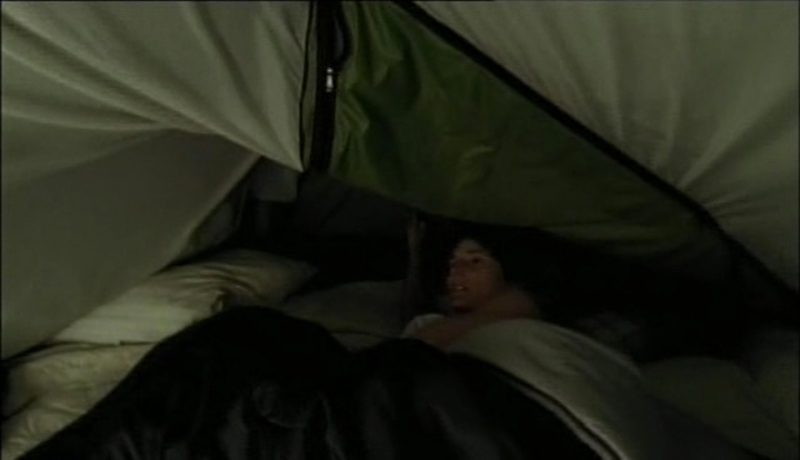 A woman with short brain hair is in a sleeping bag in a tent that appears to be caving in. She looks up in fright.