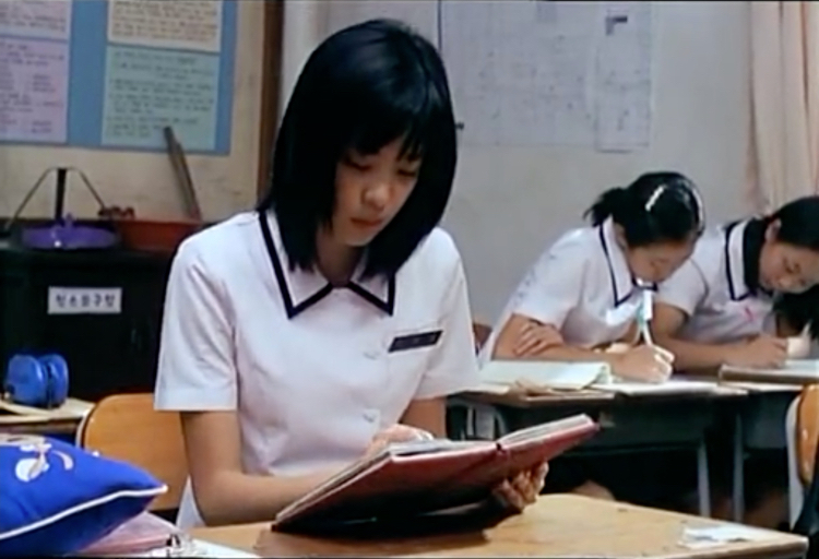 A teenage girl in a white school uniform reads a red journal while other girls write behind her.