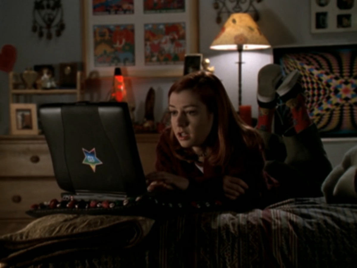 Willow lying on her stomach on her bed, using her black laptop that has a rainbow star sticker on it, with her sneakers in the air behind her