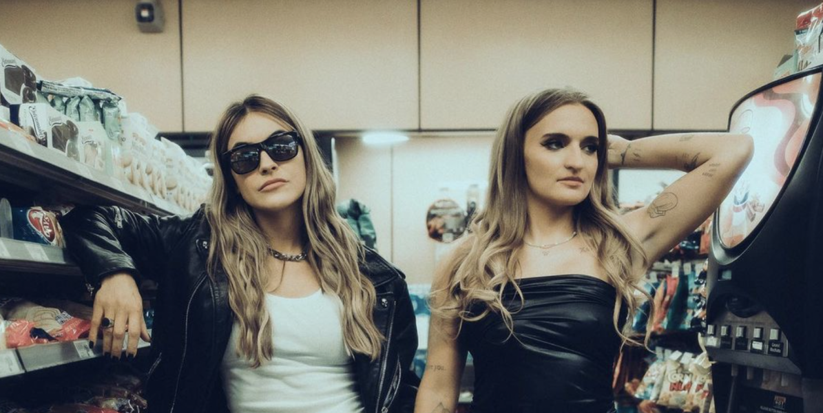Chrishell Stause and G Flip are in a convince store dressed as each other, both are white people with shoulder length blonde hair, one has on a white tank top and black leather bomber jacket with large black sunglasses, the other is in a black sleeveless mini dress