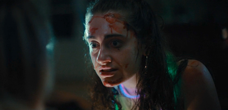 Rachel Sennott as Alice looks at someone wide-eyed with blood on her face and glow stick necklaces around her neck.