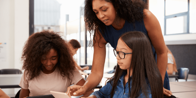 a teacher leans over two students, instructing them in the use of a tablet