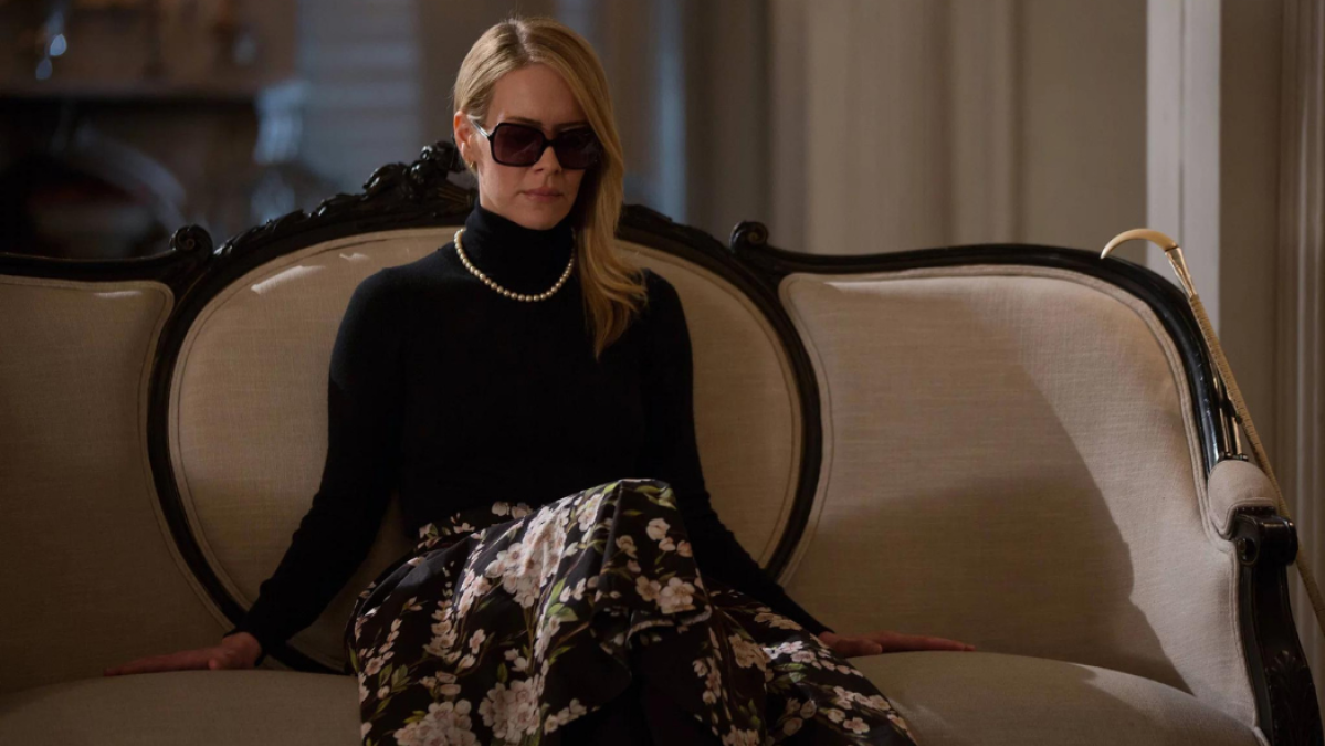 Cordelia from AHS: Coven sits on a couch wearing a black turtleneck and sunglasses