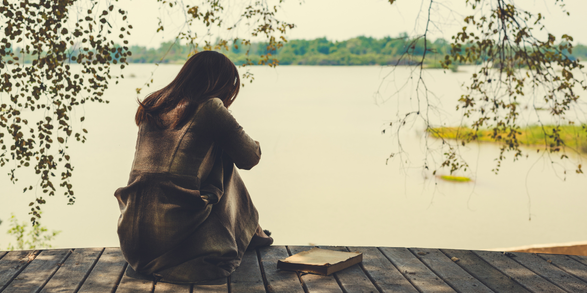 A woman with long, brown hair wears a long, brown coat. She sits on a dock overlooking a lake and hugs her knees to her chest, facing away from the camera. A book sits on the dock beside her. Tree branches hang on either side of her.