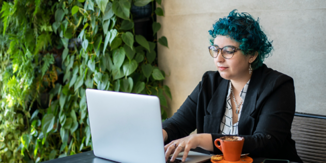 A blue haired queer person sits at a computer, and a green leaf wall is behind them