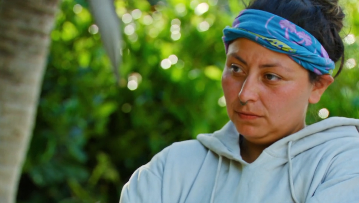 Karla Cruz Godoy, a contestant in Survivor Season 43, wears a grey hoodie and blue buff representing her membership in the Coco tribe, and looks to her right