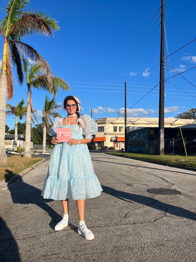 Kayla, a South Asian woman stands in a blue doll dress, holding her book, Helen House.