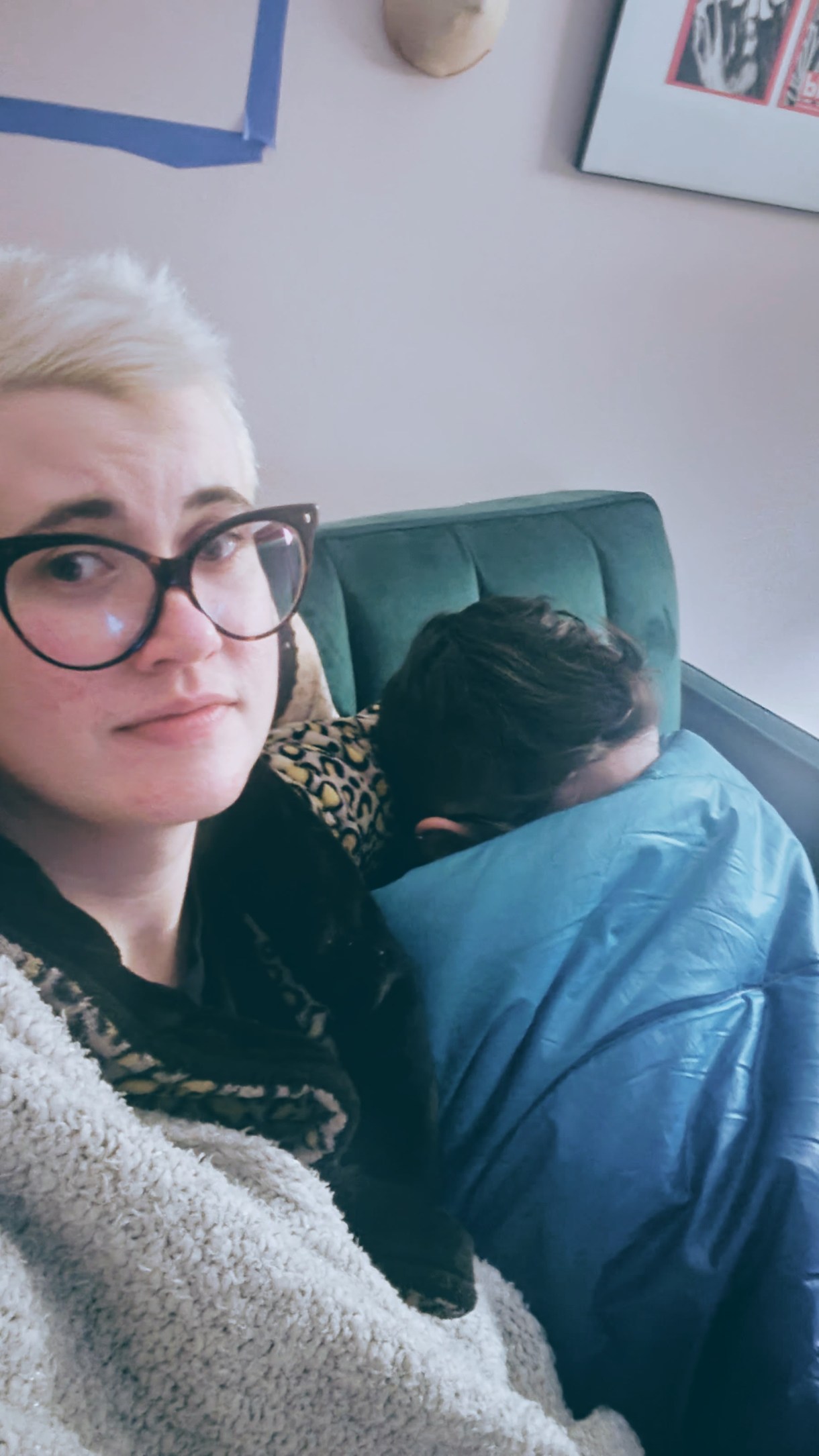 Nico, a white genderqueer human with large glasses, holds their girlfriend who is ducking beneath a blanket while watching a scary movie