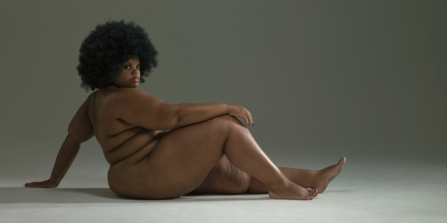 A Black model poses nude with her arm on her knee while sitting and looking at the camera.