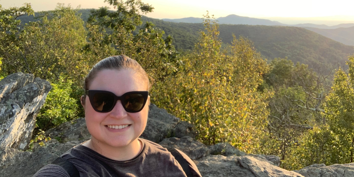 The writer Katie Reilly stands on a hiking trail with sunglasses. There are trees and mountains behind her.