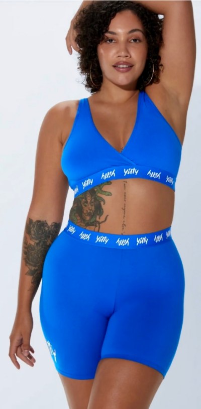 A brown skin woman with tattoos and curly hair wears a bright blue Yitty spandex bralette and biker shorts set. She faces the camera directly, with one arm propped behind her head.