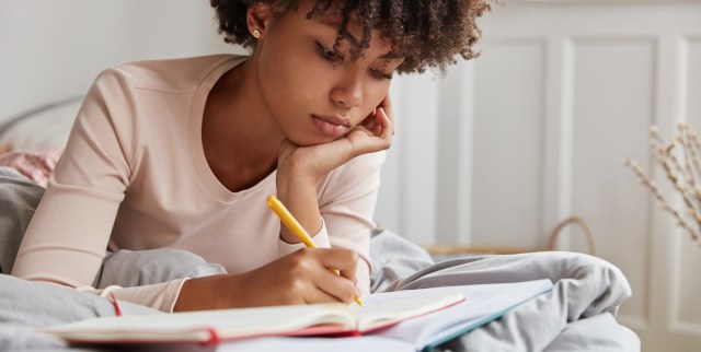 a young Black woman with short curly hair looks pensive as she writes in her journal