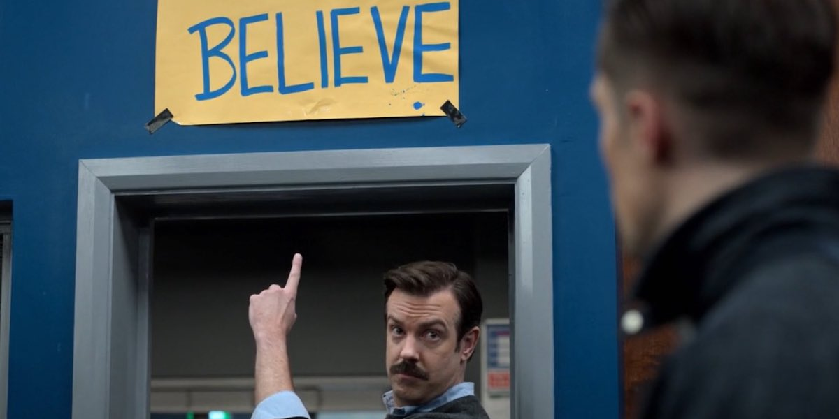 Ted Lasso pointing to the BELIEVE sign in the locker room