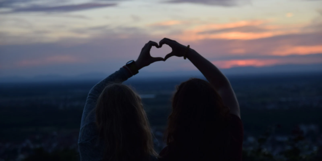 A silhouette of two teens at sunset, making a heart with their combined hands