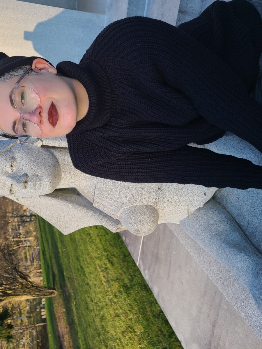 Nico, a white genderqueer human with blonde hair leans against a sphynx, wearing a black turtleneck and beanie as well as glasses and burgundy lipstick