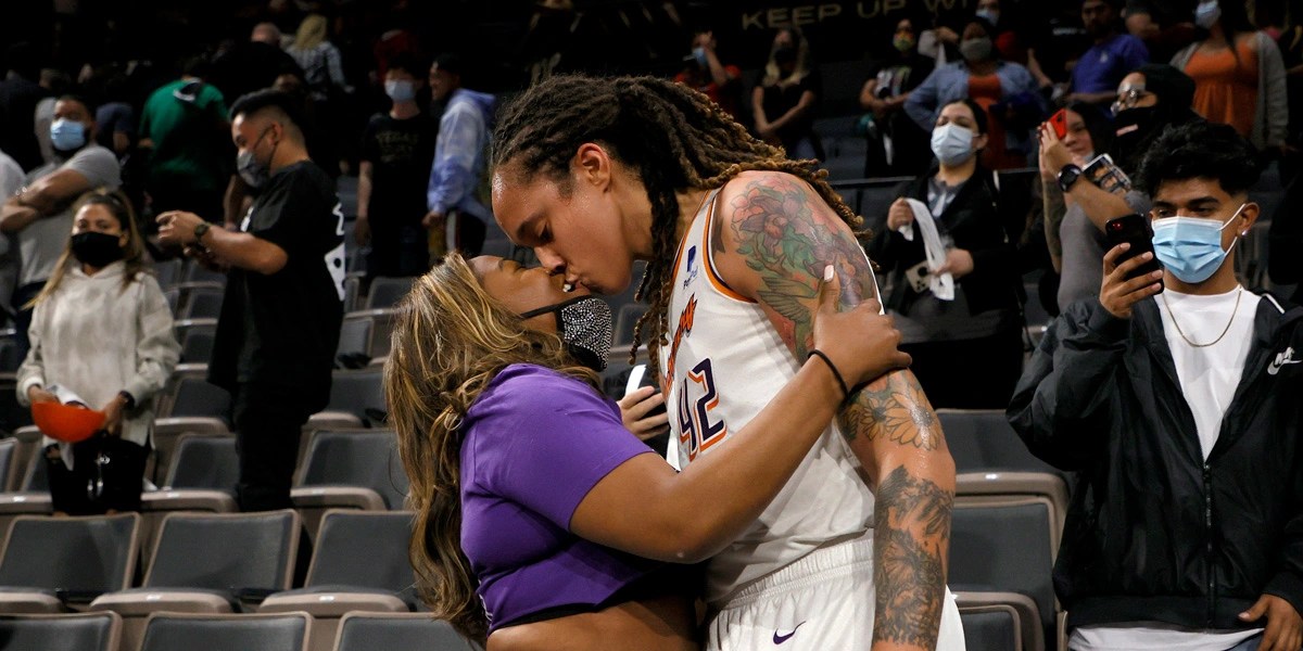 Brittney Griner kisses her wife Cherelle couurtside after a Phoenix Mercury game. Cherelle is in a purple t-shirt and Brittney is in her uniform.