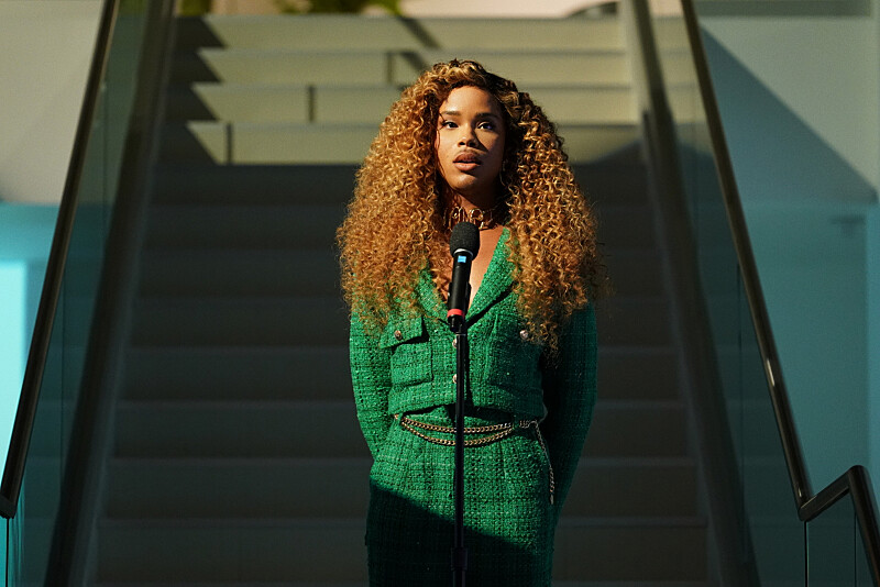 Nate stands on the stairs with her curly locks down on her shoulders. She is wearing a green tweed cropped jacket with matching pants.