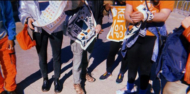 A group of queer folks are shown holding bandannas from REI. Everyone is pictured from the waist down.
