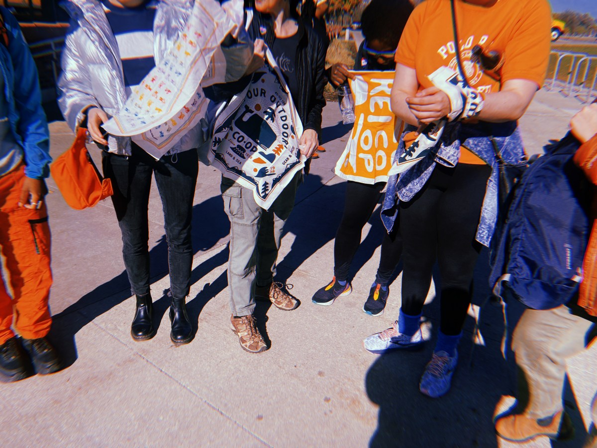 A group of queer folks are shown holding bandannas from REI. Everyone is pictured from the waist down.