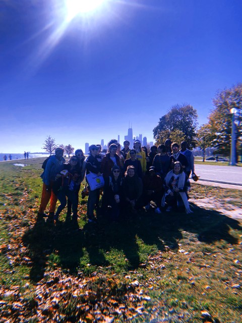 A group of queer folks huddled together smiling, while the Chicago lakefront is behind them on a sunny day.