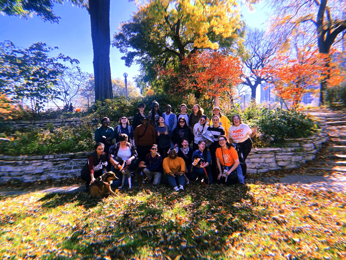 A group of queers huddled together in a park on an autumn day posing and smiling for a photo.