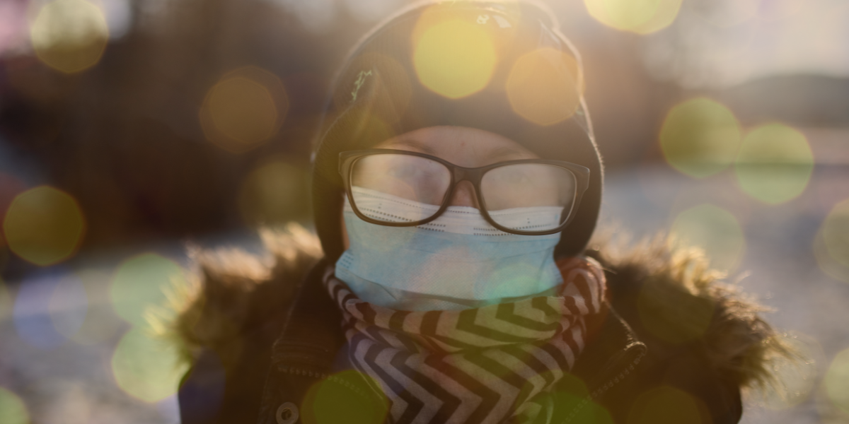A woman wears glasses and a mask as well as a beanie and winter coat and is outside in the cold