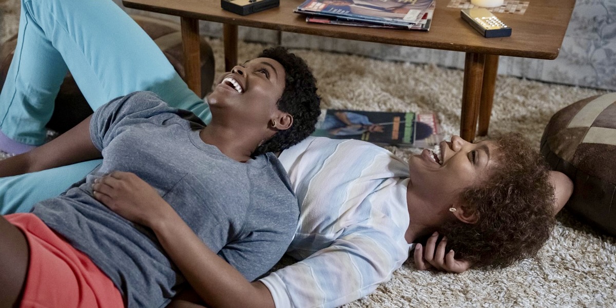In a still from "I Wanna Dance with Somebody" Naomi Ackie and Nafessa Williams are dressed in 1980s clothing with short wigs, they are laying together on the floor