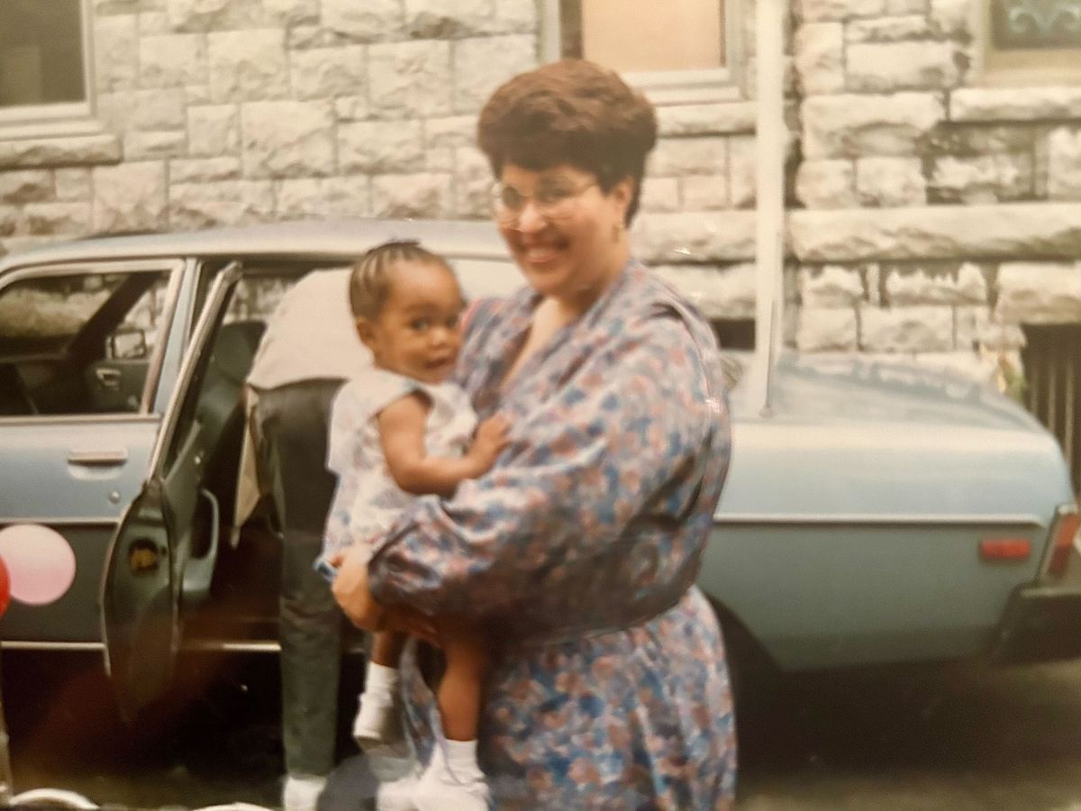 Carmen at roughly one-year-old, in her mother's arms in the late 1980s. They are standing in front of a blue cary. Her mother is wearing a blue dress.