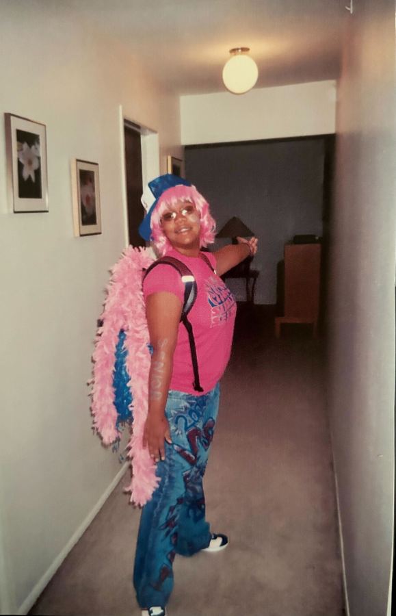 Carmen, a Black femme, in her senior year of high school, dressed for spirit week in a pink wig, sunglasses, a blue trucker hat, decorated and drawn on blue jeans, a pink tee shirt and a pink feather boa, stands in her hallway, arm stretched out to the side