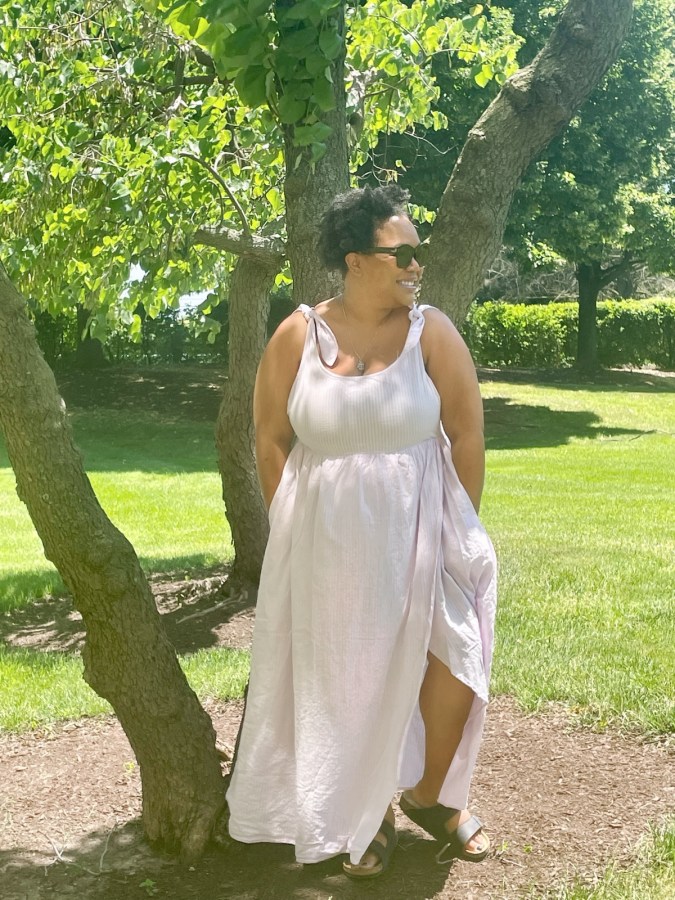 Carmen is in a flouncy purple dress with a slit and black Birkenstock sandals and black sunglasses. She's standing in front of a tree on a bright sunny day.