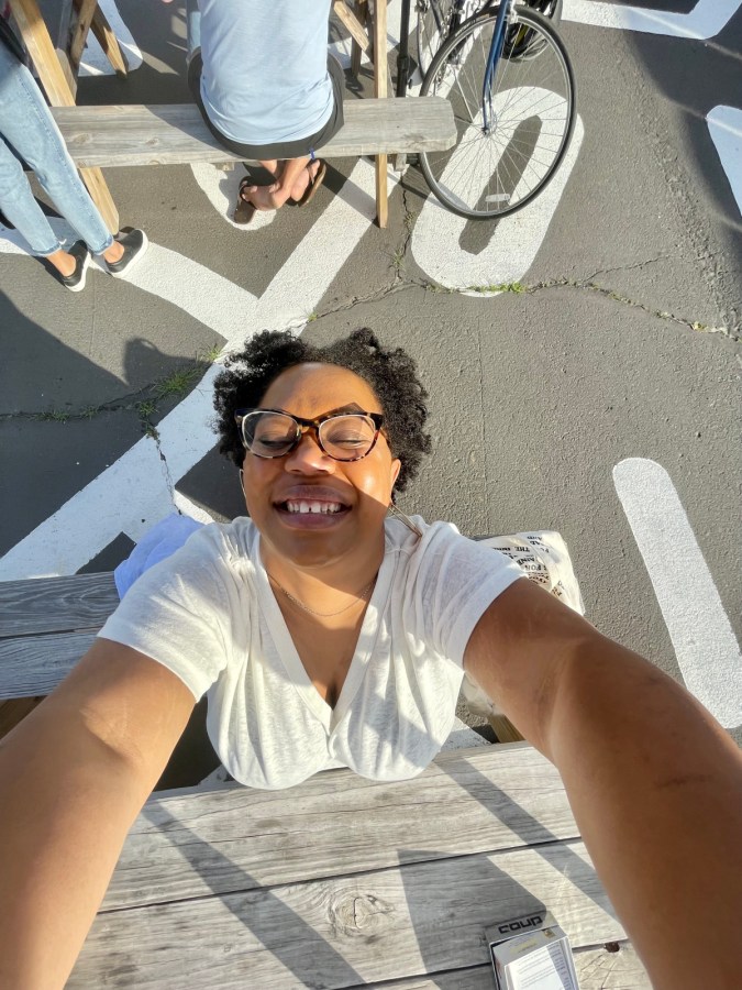 Carmen is smiling upside down taking a selfie outside at a picnic table in a parking lot.