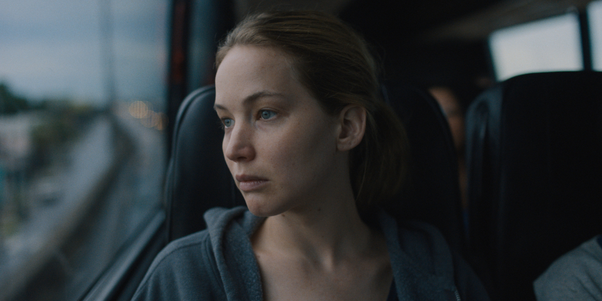 Jennifer Lawrence stares out a bus window in a hooded sweatshirt and her hair pulled back.