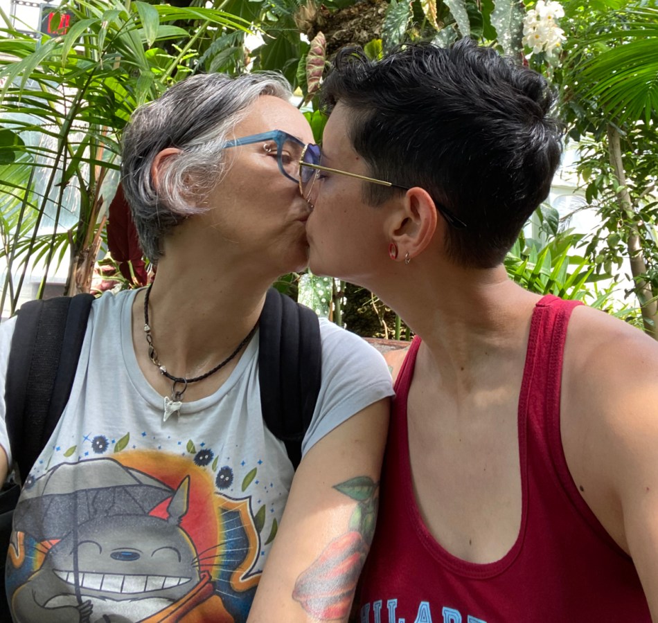 Tracy and Mia kiss in some kind of garden. This is a recent photo. Mia's tattoo of a pepper is visible and she is wearing a totoro tee shirt. tracy is weraing a red tank. mia's hair is gray and tracy's is black and they both wear glasses.
