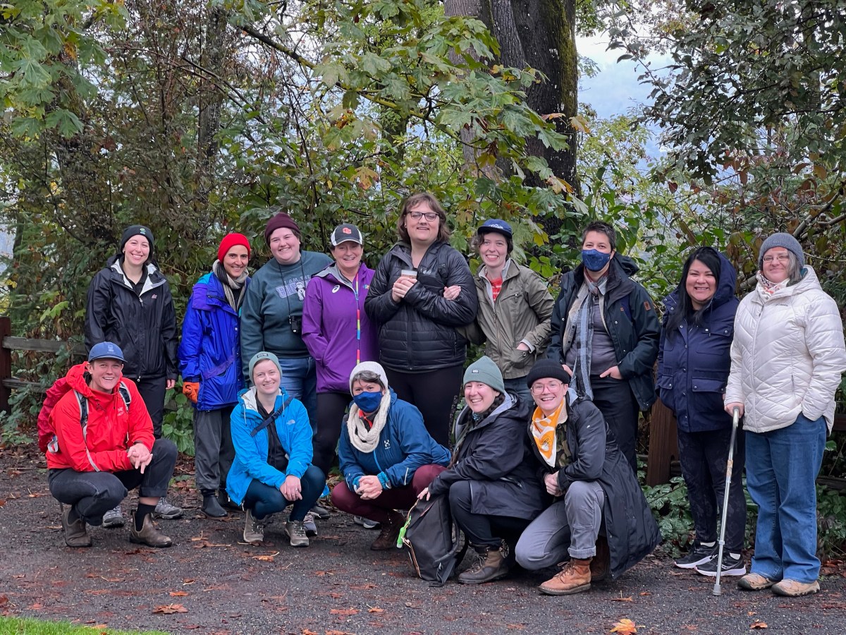 14 queer people posing for a group photo before embarking on a hike. Some squat, some stand. A few wear masks. Everyone is dressed and ready for rainy weather.