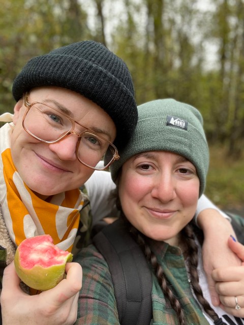 Two dykes take a selfie — one wears an REI bandana and chomps on a green apple with pink insides, the Mountain Rose apple, and the other wears a green REI beanie over her pigtail braids.