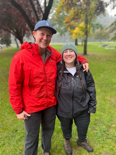Two queers, Sarah from REI and Vanessa from Autostraddle, pose at the trailhead. Sarah wears a bright red rain jacket and an enormous smile. Vanessa wears a black rain jacket and an equally huge smile.