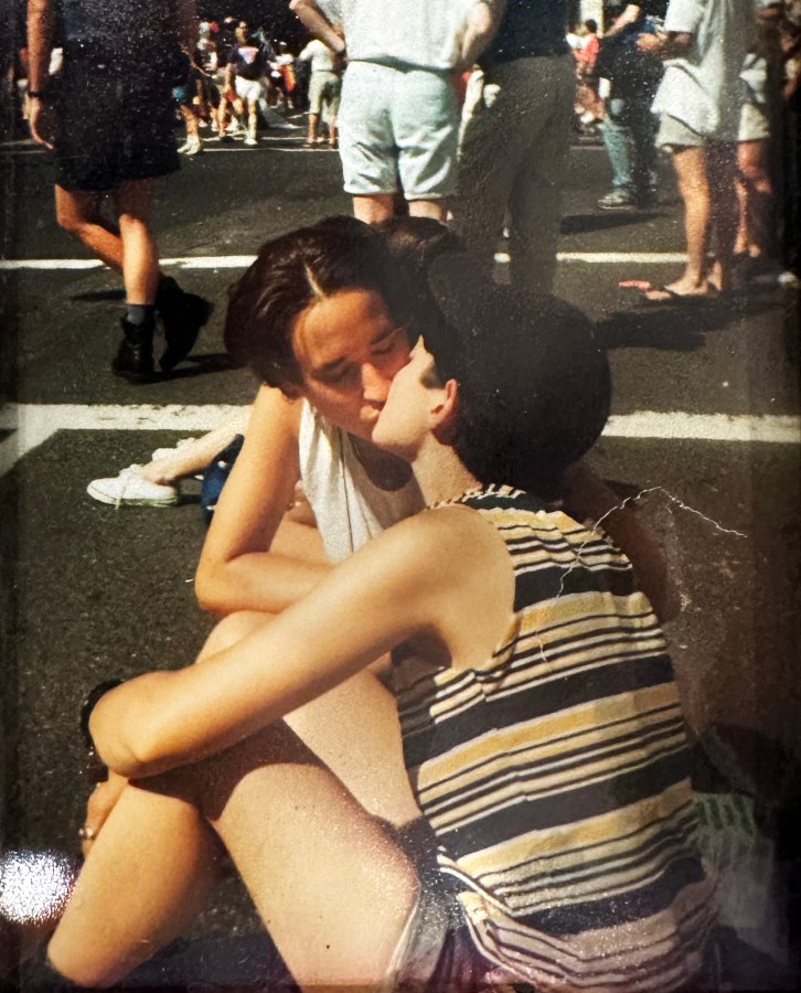 Tracy and Mia, in this older photo, sit on the asphalt at a Pride event and kiss. They both have short hair and are wearing tank tops and shorts. Tracy has on a baseball cap.